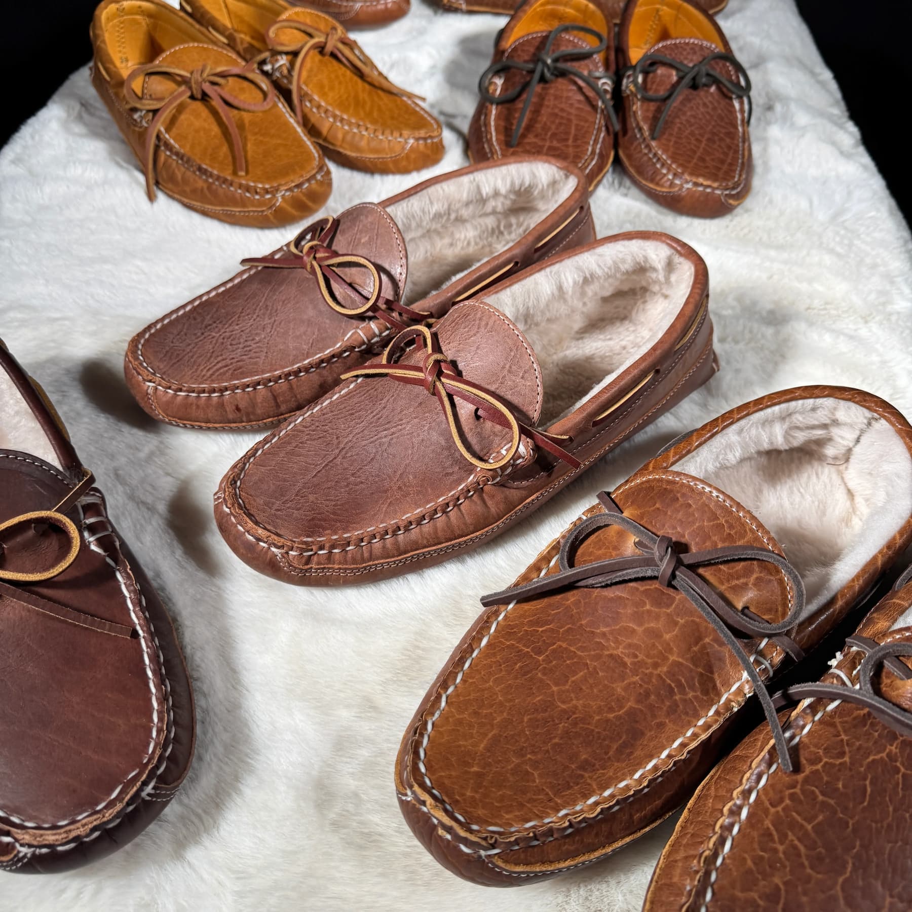 Collection of handcrafted leather moccasin slippers in various brown shades displayed on a soft white sheepskin surface.