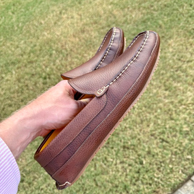 Brown leather loafers held by a hand with a grassy background