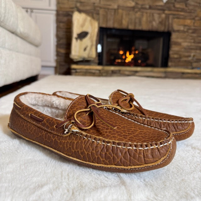 Pair of brown moccasin slippers on a white surface with a fireplace in the background.