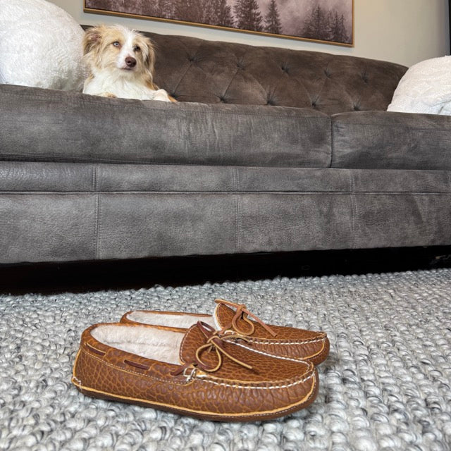 Brown moccasin slippers on a textured rug with a gray sofa and dog in the background.