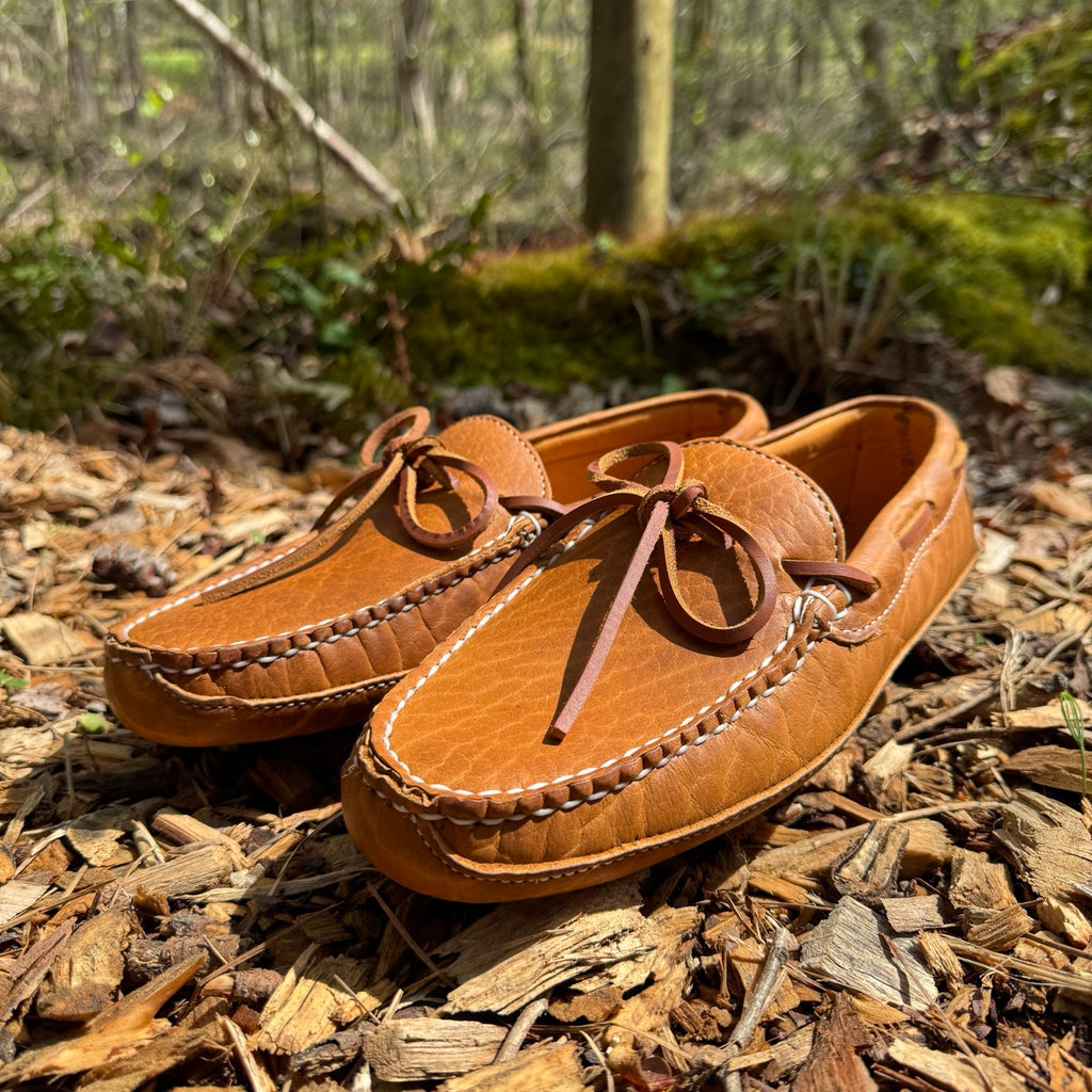 Pair of brown leather moccasins on a forest floor with greenery in the background