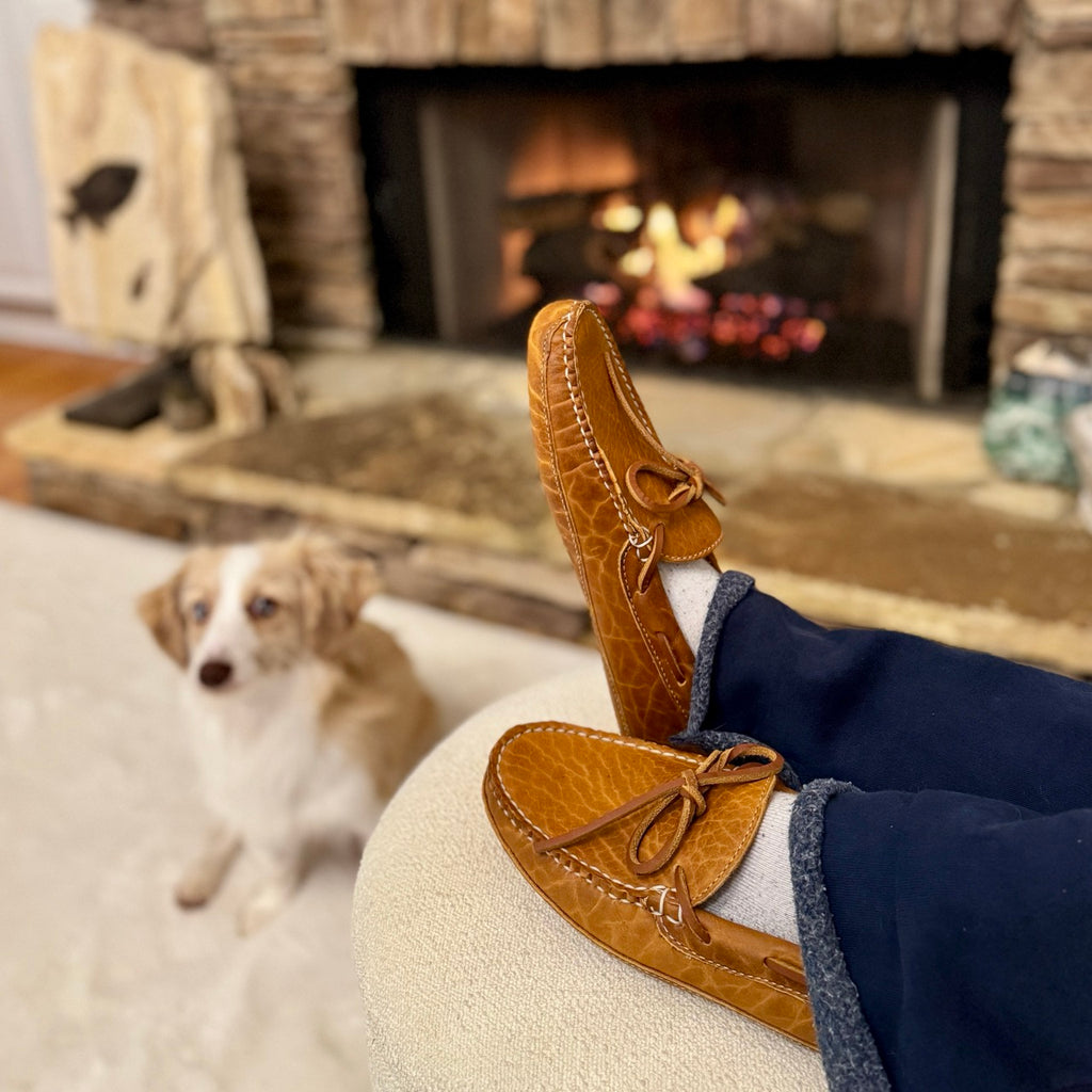 Person wearing brown moccasins with a dog sitting on a couch in front of a fireplace.
