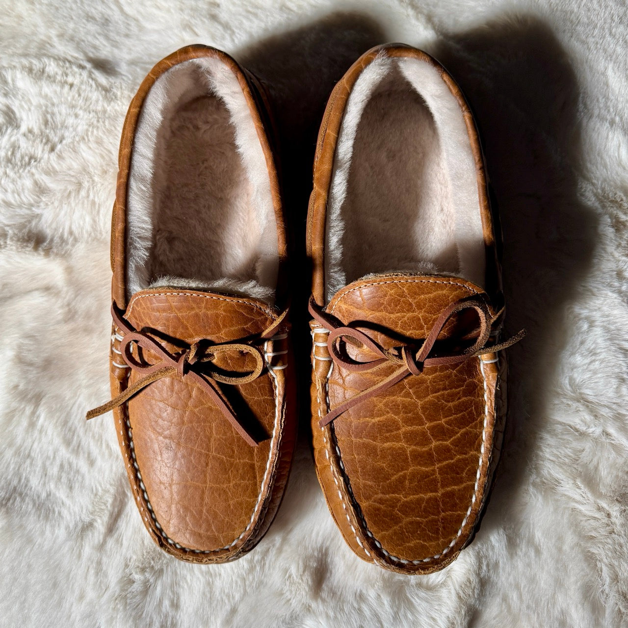 Pair of brown leather moccasin slippers with fur lining on a textured white surface.