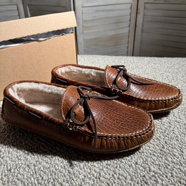 Pair of brown moccasin slippers with fur lining on a textured surface.
