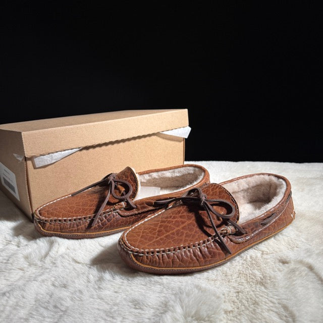 Brown leather slippers with fur lining on a white surface next to a cardboard box.