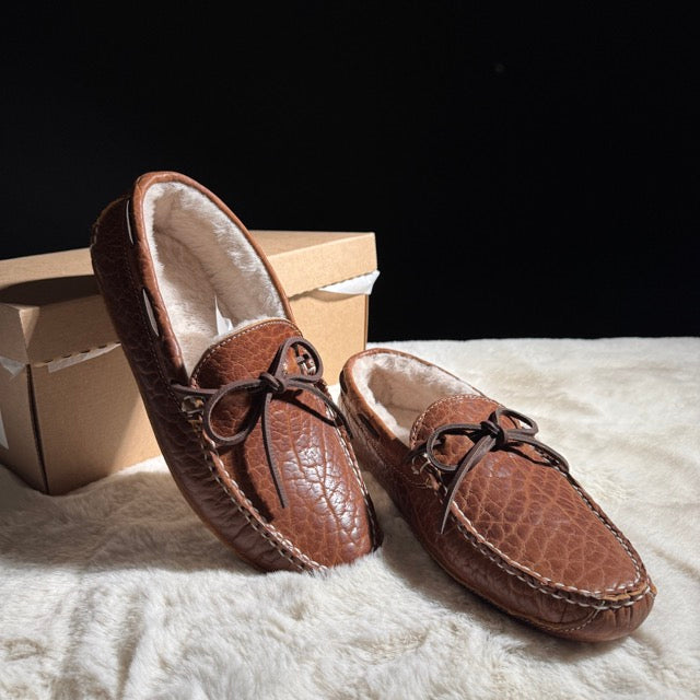 Pair of brown leather moccasin slippers on a white surface with a cardboard box in the background.